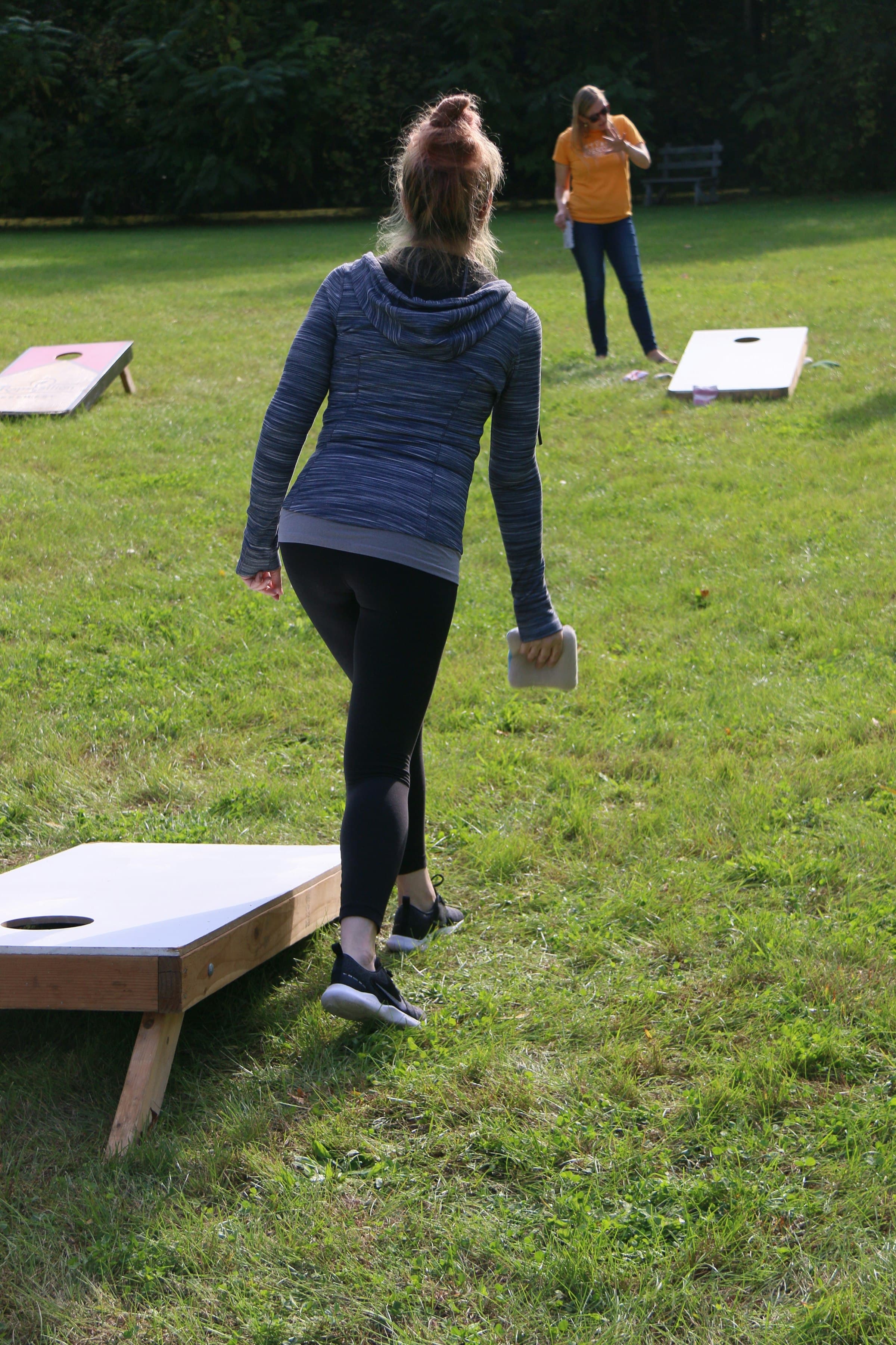 Two players mid-throw at an outdoor cornhole tournament on a grassy lawn.